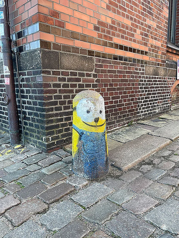 Gelber, bemalter Poller mit Gesicht und blauer Kleidung vor einer Backsteinwand auf einem gepflasterten Weg.