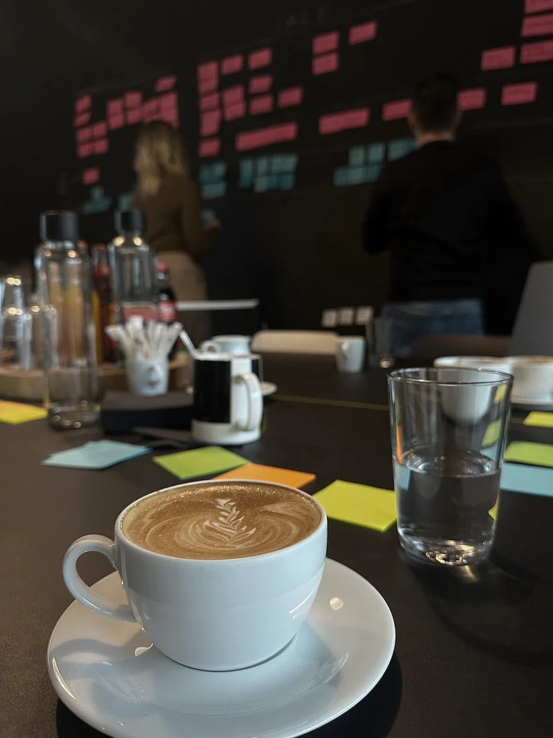 Tasse mit Latte Art auf einem Tisch mit Glas Wasser und bunten Notizzetteln, im Hintergrund zwei Personen vor einer Wand mit vielen farbigen Haftnotizen