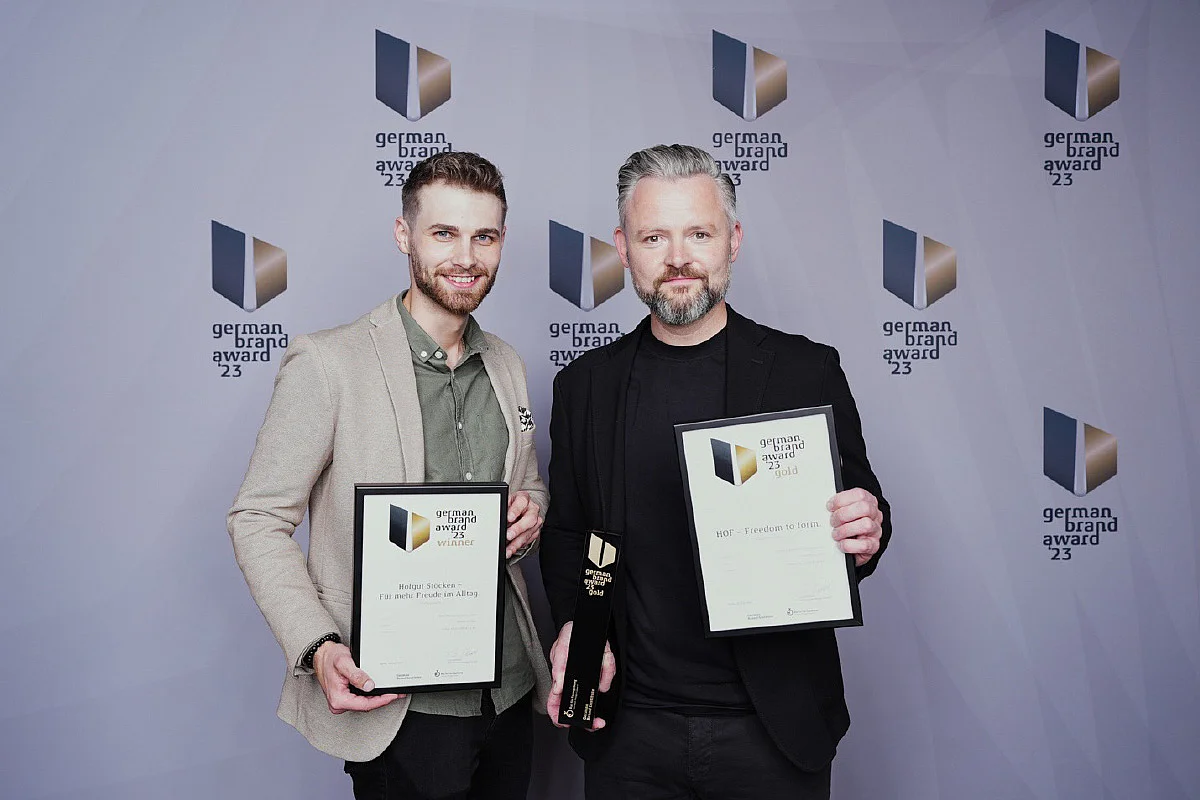 Zwei Männer halten Auszeichnungen vor einer Hintergrundgrafik mit dem Text „German Brand Award 23“.