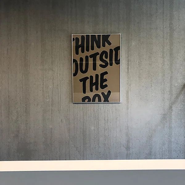 Ein gerahmtes Poster mit dem Text "THINK OUTSIDE THE BOX" an einer Betonwand.