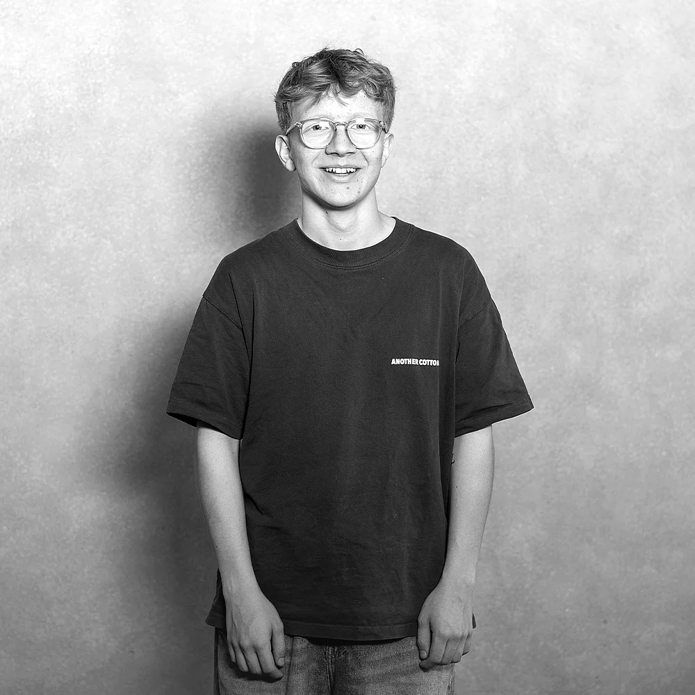 Ein junger Mann mit Brille trägt ein T-Shirt und steht vor einem hellen, einfarbigen Hintergrund.