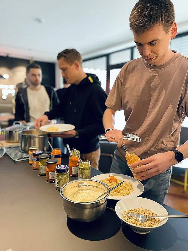 Drei Personen bereiten Essen in einer modernen Küche mit Nudeln, Saucen, Gemüse und Nüssen vor.