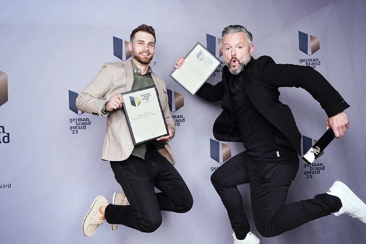 Zwei Männer springen mit Auszeichnungen vor einer Wand mit Aufschrift „German Brand Award 23“.