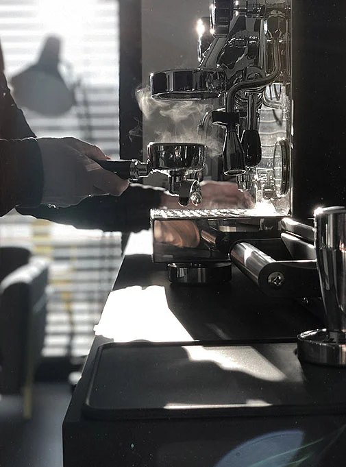 Eine Hand hält einen Siebträger vor einer Kaffeemaschine, aus der Dampf aufsteigt.