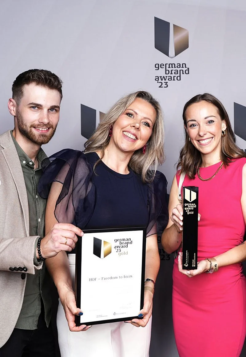 Drei Personen halten eine Urkunde und einen Preis vor einem Hintergrund mit dem Logo "German Brand Award 23".