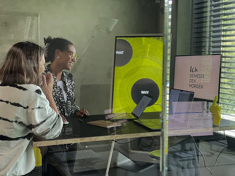 Zwei Personen stehen an einem Tisch mit Laptops und Monitoren in einem modernen Raum. Bildschirm zeigt Text.