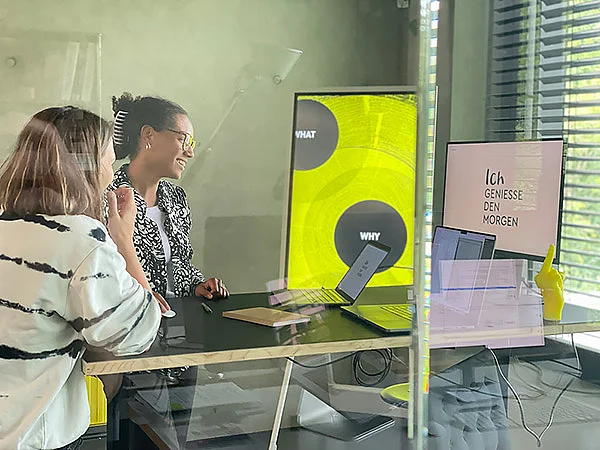 Zwei Frauen sitzen an einem Tisch mit Laptops und Monitore, umgeben von Glaswänden in einem modernen Raum.