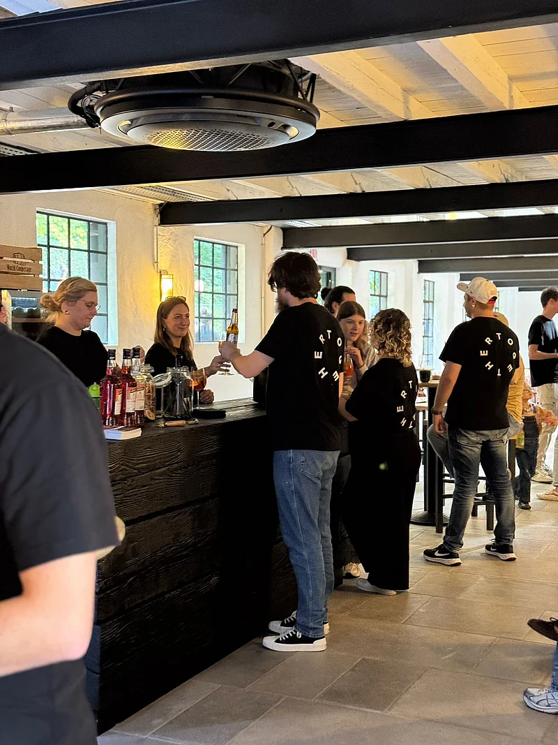 Eine Bar mit Menschen in schwarzen T-Shirts, die Getränke servieren und konsumieren. Tische und Fenster sind sichtbar.