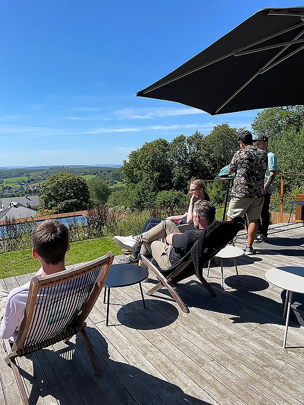 Personen sitzen und stehen auf einer Holzterrasse mit Blick auf eine grüne Landschaft und Himmel. Sonnenschirm und Möbel vorhanden.